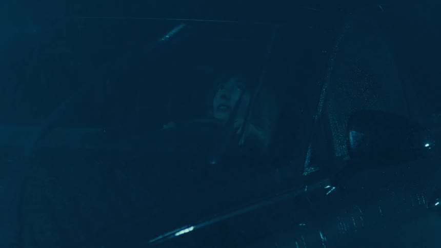 A closeup of a woman sitting in the drivers seat gazing through the windshield as heavy rain pours outside at night