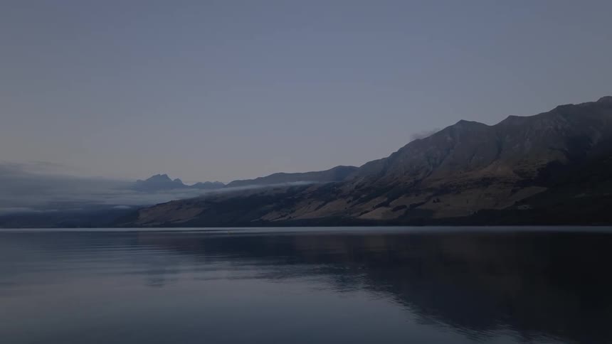 A still dark lake reflects imposing shadowy mountains under a dim evening sky