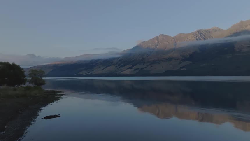 A calm lake stretches along a rocky shoreline towards a backdrop of mountains partially obscured by low lying clouds under a dusky sky