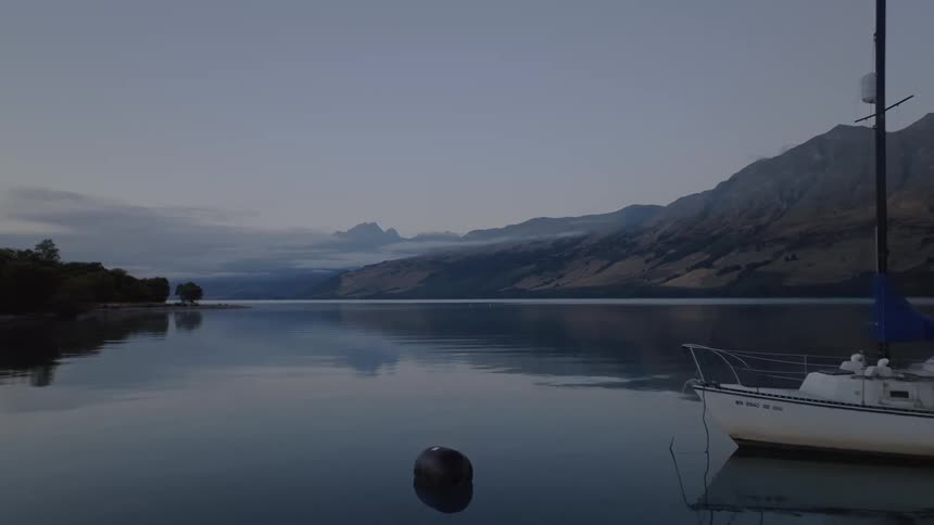 A sailboat with a blue sail cover is anchored on a calm lake surrounded by mountains and trees under a clear sky