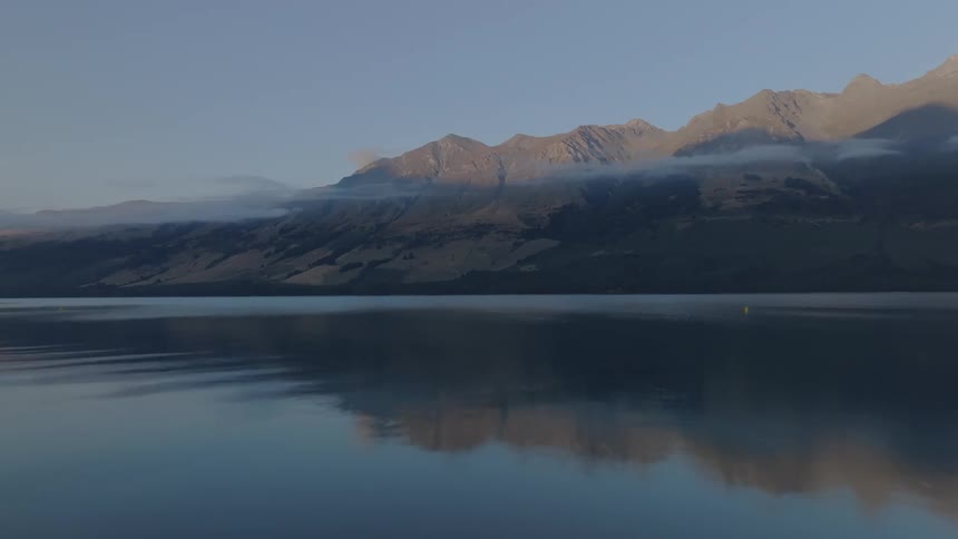 A calm dark lake extends toward a range of rugged mountains under a clear early morning or late evening sky