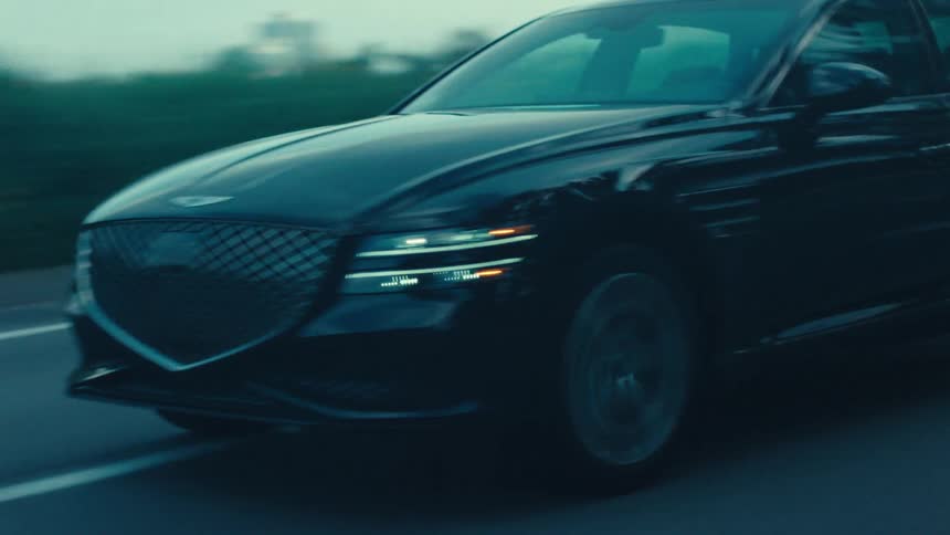 A darkcolored genesis sedan drives on a paved road lined with trees and residential buildings under a muted sky