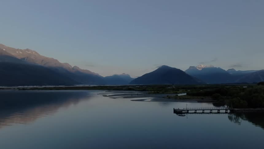 A slow aerial pan reveals a serene mountain lake reflecting the peaks at dawn or dusk moving past a lone sailboat to a pier and braided river delta