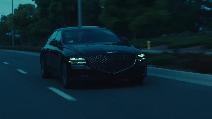 A sleek black genesis sedan cruises down a dimly lit road at dusk with its distinctive headlights on