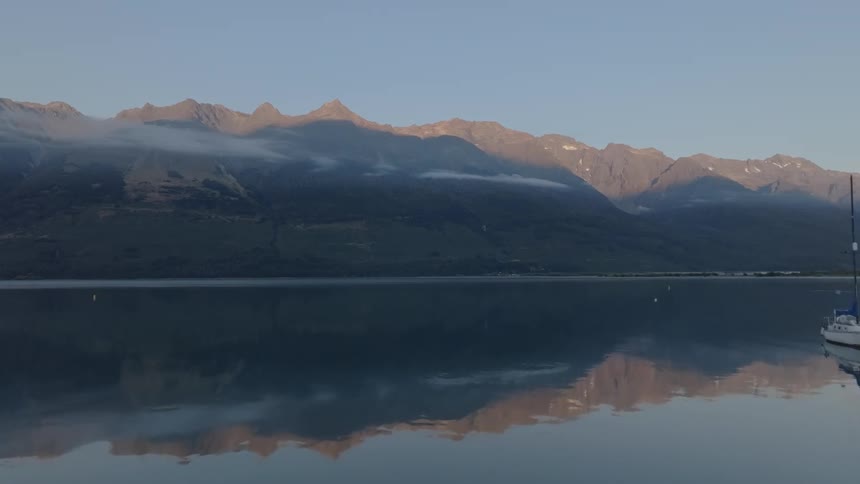 A lone sailboat rests on a calm lake reflecting the silhouettes of a mountain range under a twilight or morning sky