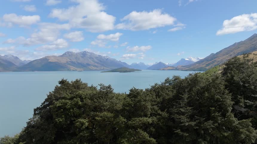 A wide pale blue lake stretches towards a backdrop of rugged mountains under a partly cloudy sky framed by dark green trees in the foreground