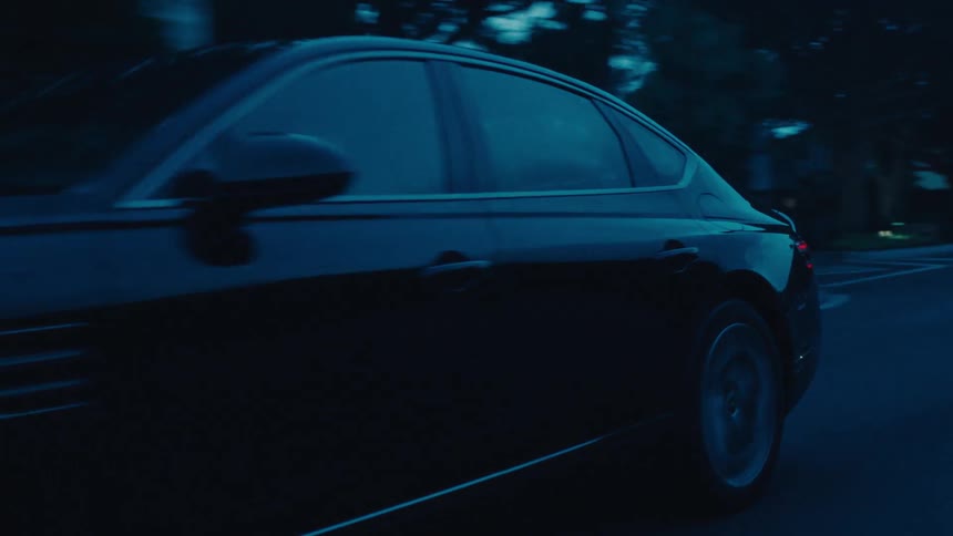 A dark colored genesis sedan drives on a paved road lined with trees and residential buildings under a muted sky