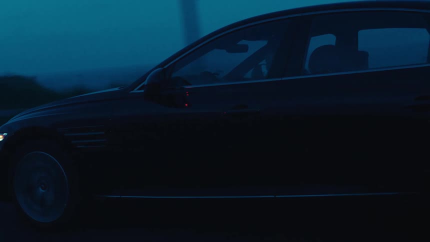 A black sedan travels along a dark road during dusk or dawn near the ocean