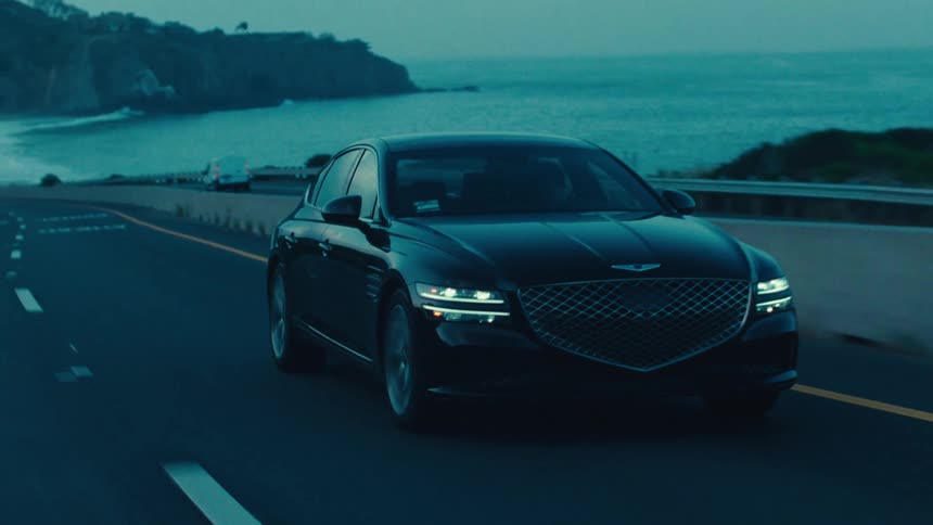 A black genesis sedan drives along a coastal highway at dusk with the ocean visible in the background