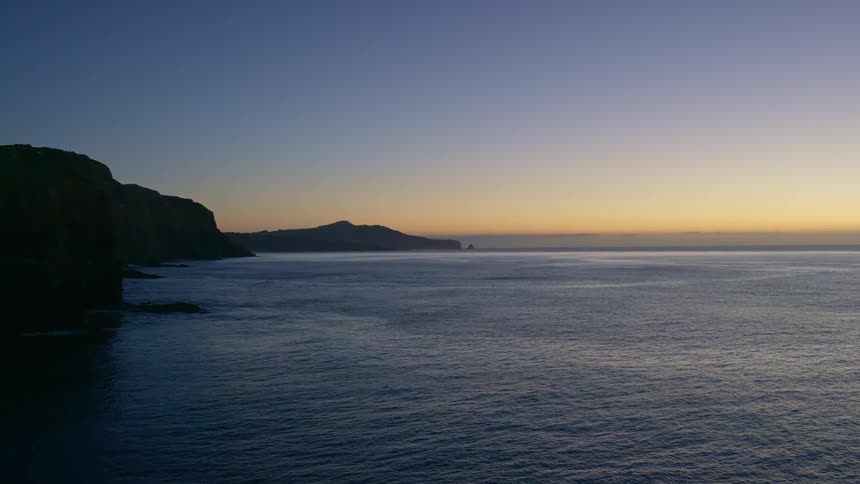 A dark rocky coastline meets a vast undulating ocean under a gradient sky transitioning from deep blue to a soft orange glow on the horizon