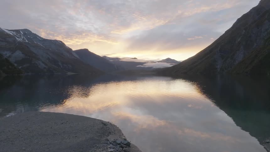 A still lake reflects the light of the early morning or late evening sky surrounded by steep dark mountains
