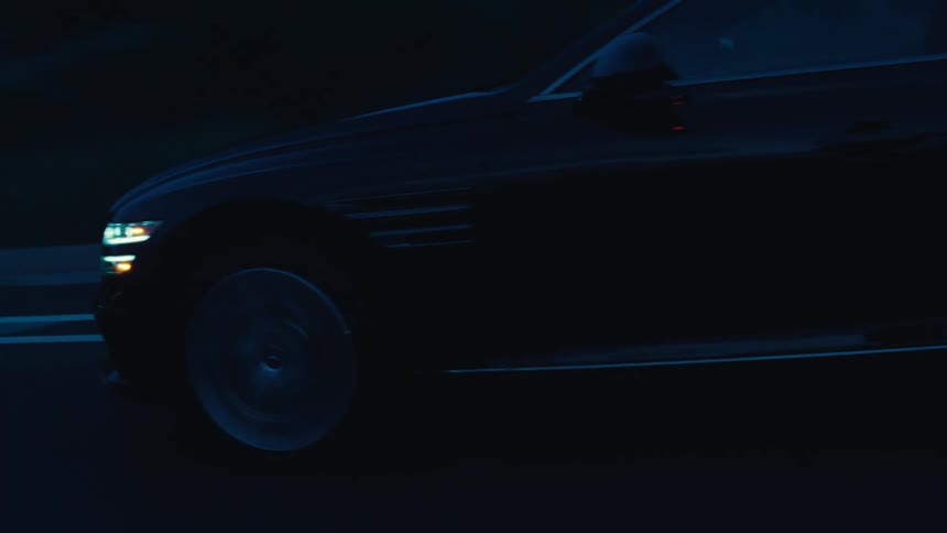 A black sedan moves along a dimly lit coastal road during dusk or dawn