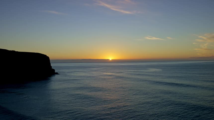 The sun sets on the horizon over a wide rippled expanse of ocean adjacent to a dark rocky landmass