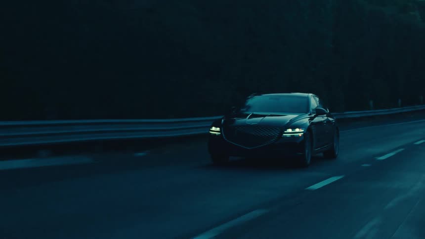 A black sedan travels along a dark road during dusk or dawn