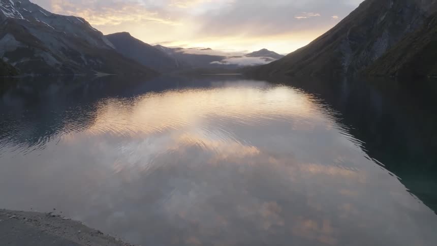 A tranquil lake reflects the soft glow of the sky at dusk nestled between steep rocky mountains with patches of snow