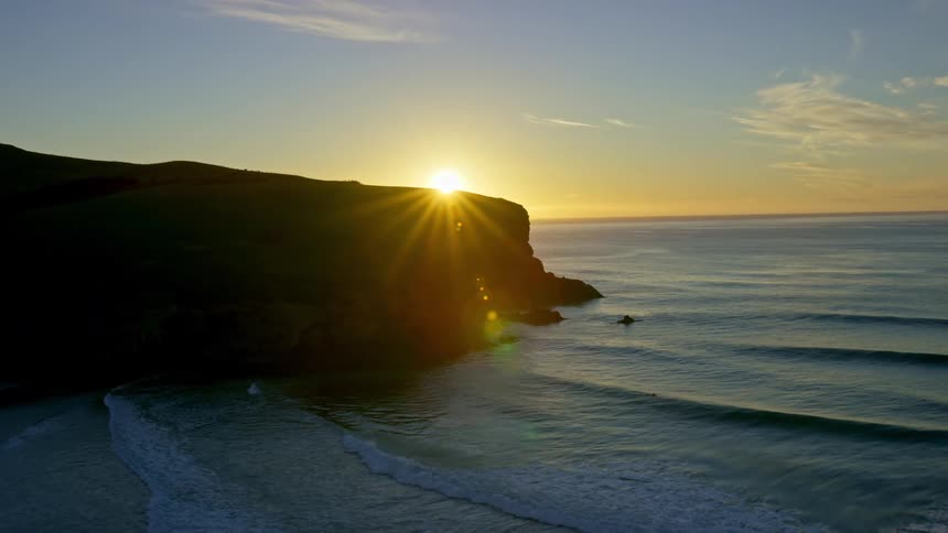 The sun rises or sets behind a dark cliff overlooking a wavy ocean under a clear sky with a few scattered clouds