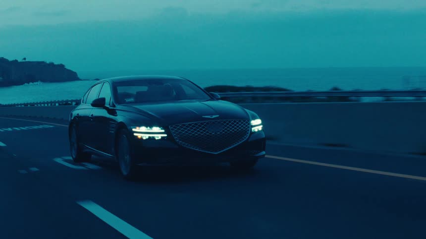 A black sedan moves along a coastal road during dusk or dawn