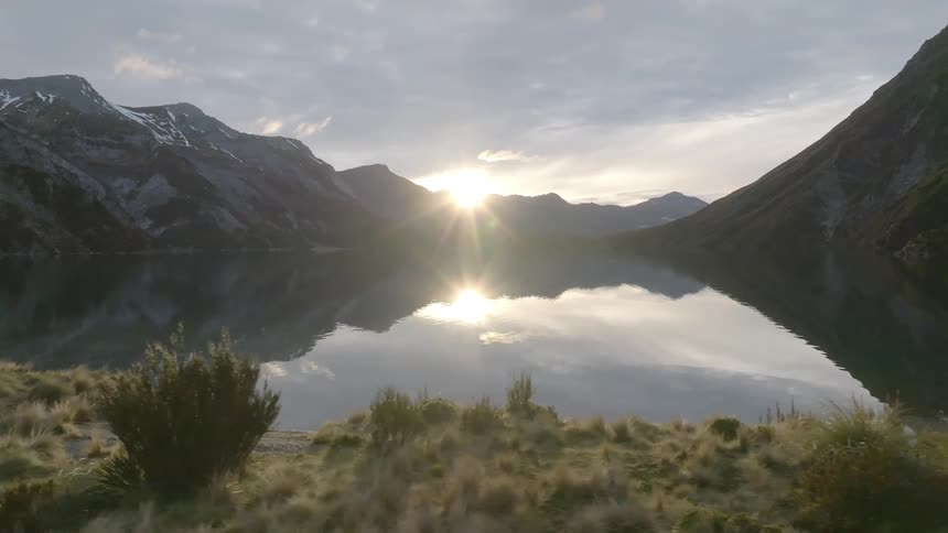 The sun rises over a tranquil lake nestled between steep rocky mountains casting a bright glare on the waters surface