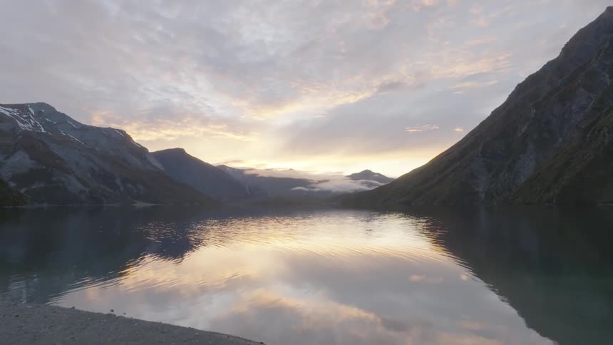 A tranquil lake reflects the soft glow of the sky at dusk nestled between steep rocky mountains with patches of snow