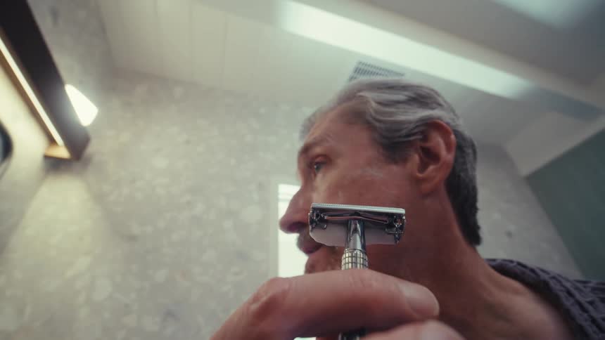 A closeup captures a man with graying hair and a partial beard shaving with a safety razor appearing to nick himself while setting the blade aside