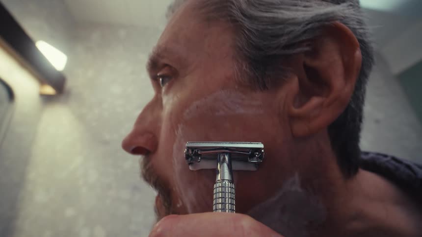 A man shaves the left side of his face while looking at the bathroom mirror