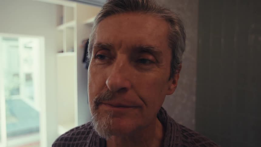 A closeup of a man with a halfshaven face admiring his work while his beard partially remains in a home bathroom