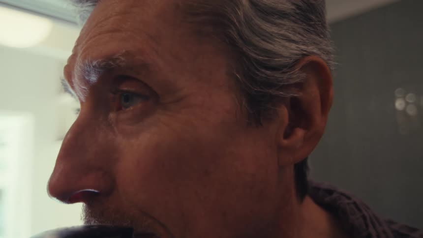 An older man uses an electric razor to shave his mustache and beard while standing in the bathroom