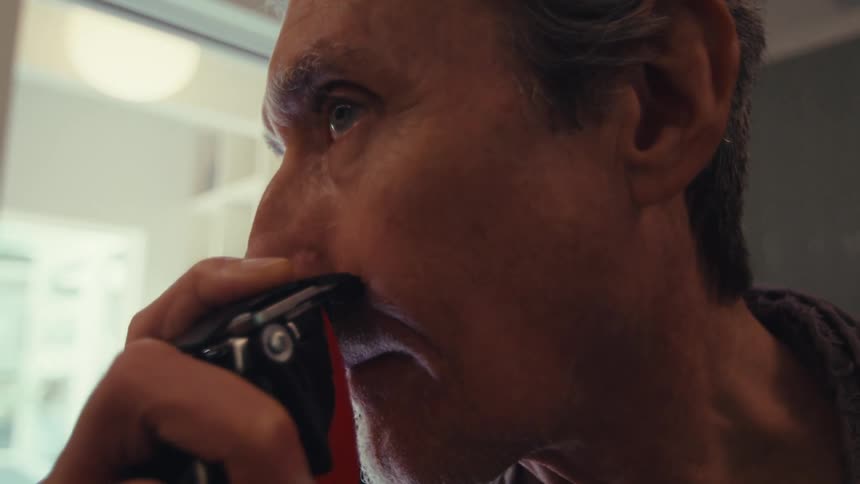 A man uses an electric shaving machine while standing near a window with light streaming into the room