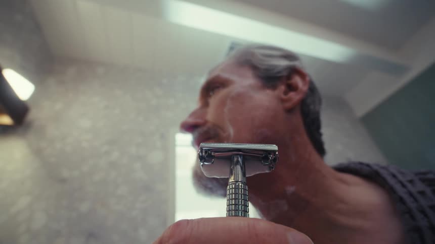 An older man shaves his neck with a manual razor while standing in the bathroom