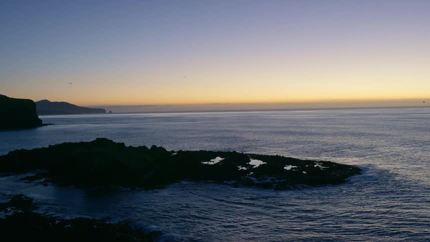 Dark jagged rocks emerge from a rippling ocean under a gradient sky transitioning from deep blue to a soft orange glow on the horizon