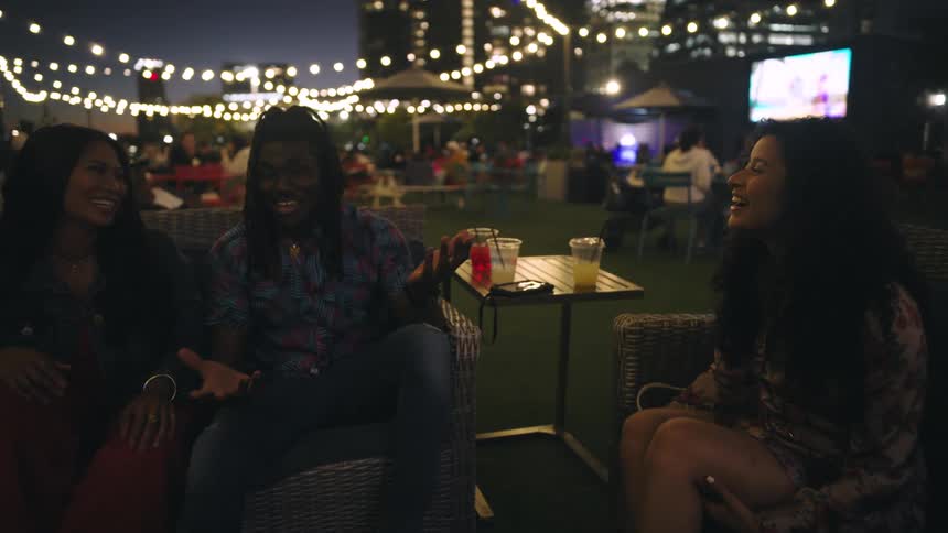A group of friends are sitting and laughing in an outdoor restaurant at night while drinking alcoholic beverages