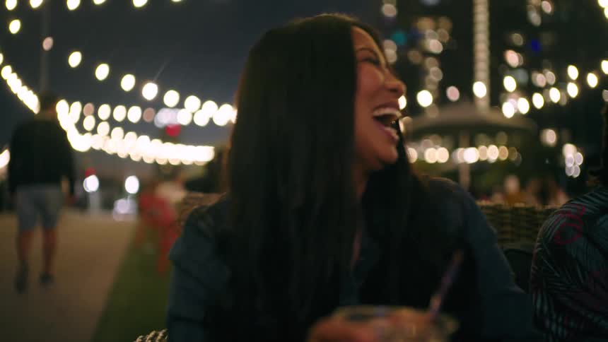 Woman holding a drink laughs with friends at an outdoor table near the waterfront at night