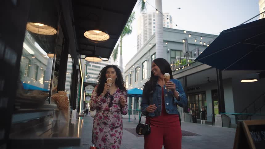 Female friends eat ice cream while walking past the outdoor counter of an ice cream shop