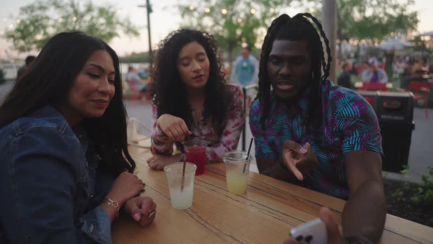 A group of friends are sitting outdoors at a picnic table while drinking and looking at a smartphone
