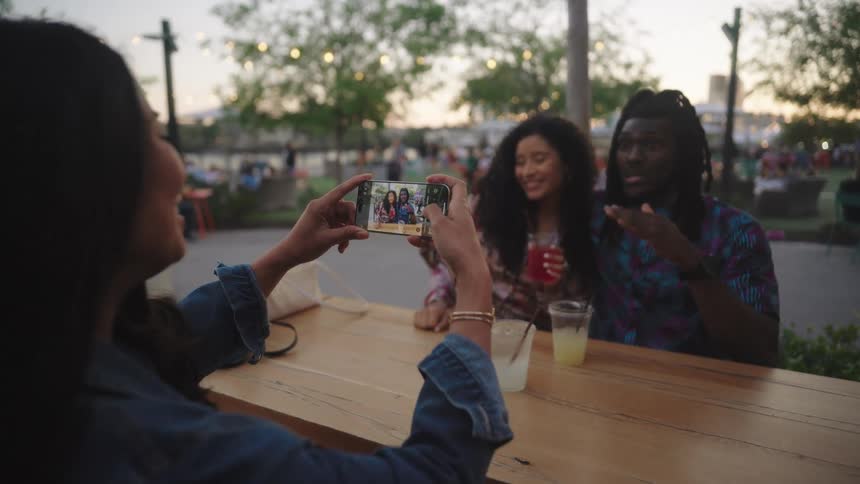 Woman takes a picture of her friends while sitting at an outdoor table at a festival after dusk