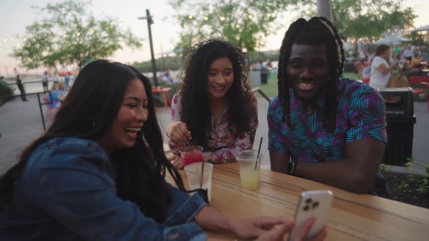 Three happy happy friends sitting at outdoor bar table talking and interacting with smartphone