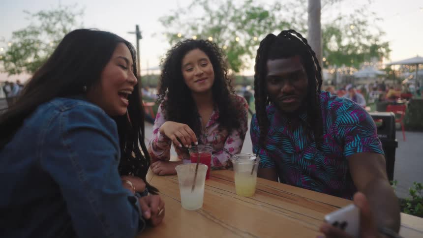 Friends sitting at an outdoor bar table laughing and interacting on a cell phone