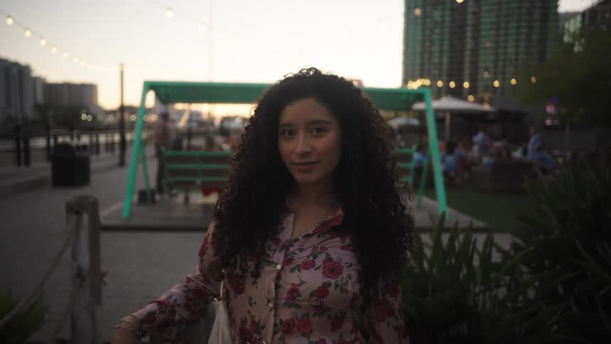 Woman standing outdoors smiles while people enjoy the waterfront park in the background after dusk