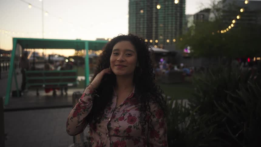 Curly haired woman with smiling facial expression at an outdoor bar with buildings in the background