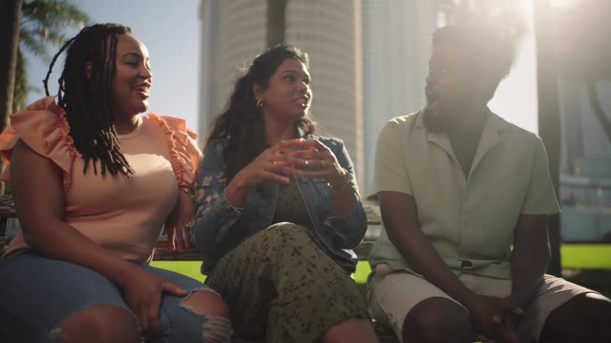 Three happy friends sitting on a bench in a square talking