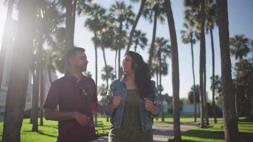 A smiling couple strolls hand in hand along a sunny path lined with palm trees enjoying a leisurely walk