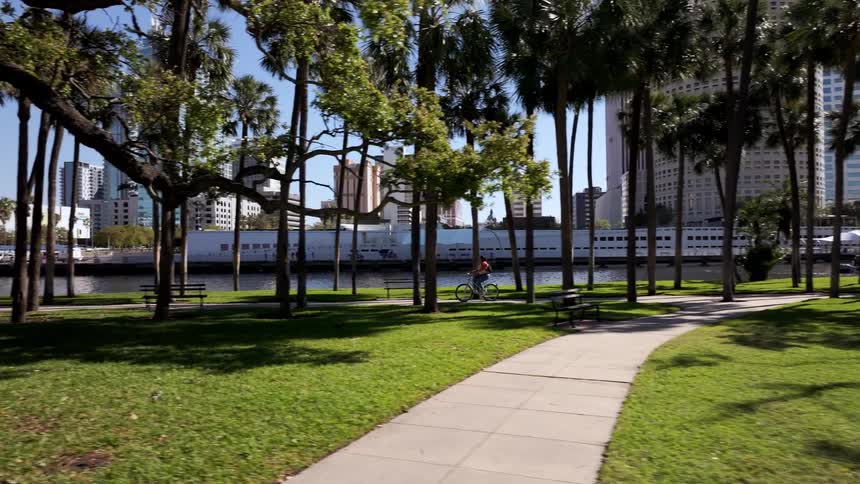 Person riding a bicycle through a treelined square with buildings in the background