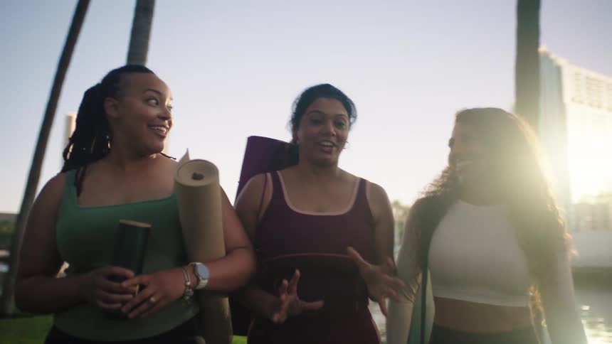 Three diverse friends in athletic gear laugh and chat while walking with yoga mats through a sunny city park near water featuring palm trees and a backlit skyline