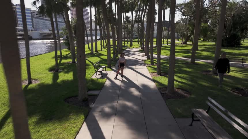 A young woman rollerblades along a riverside path framed by towering palm trees with a city skyline visible in the distance