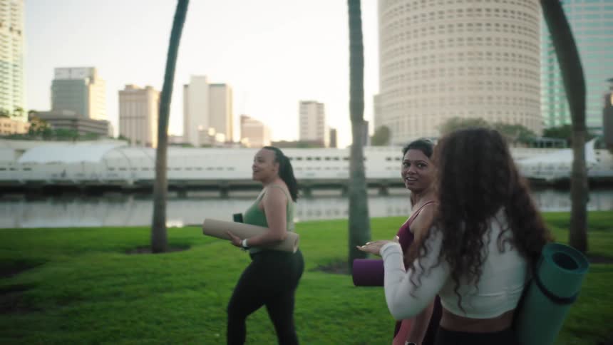 Three women carrying yoga mats walk across a lawn in an urban area with tall buildings in the background