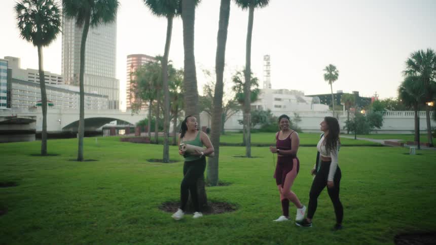Three women carry yoga mats as they walk onto the grass in a park