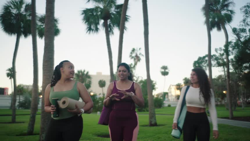 Three friends walk through a riverside park carrying yoga mats on a summer day