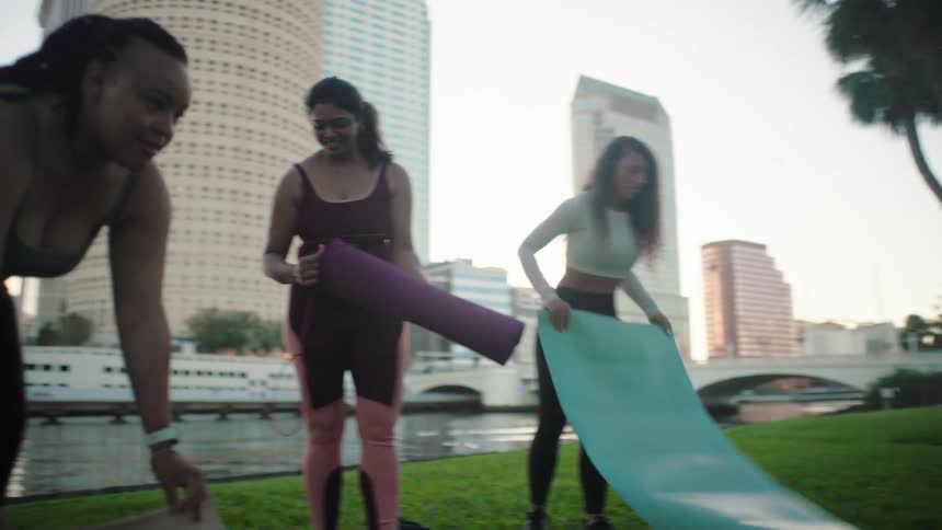 Women rolling out yoga mats on waterfront lawn in the park in the city