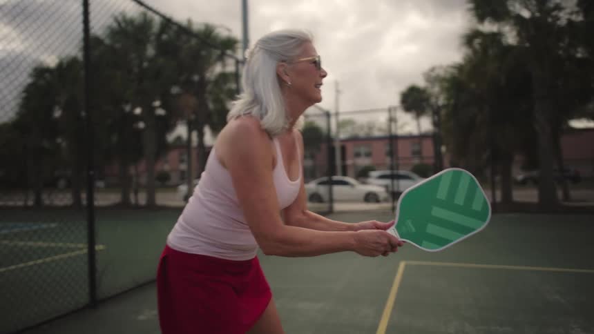 Elderly woman plays pickleball outdoors on a fencedin court under a partly cloudy sky with bright sunlight