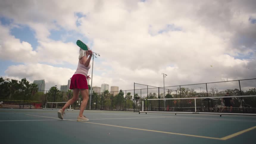 Elderly woman prepares to bat during pickleball match on outdoor court with cloudy sky and buildings in background